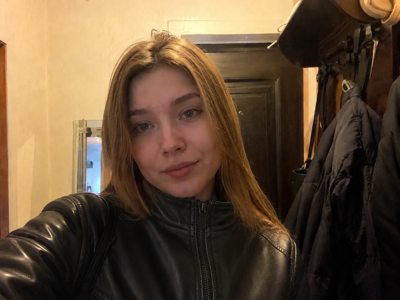Девушка в прихожей: бежевая стёганая куртка заменена на чёрную кожаную, поза, свет и фон остались теми же