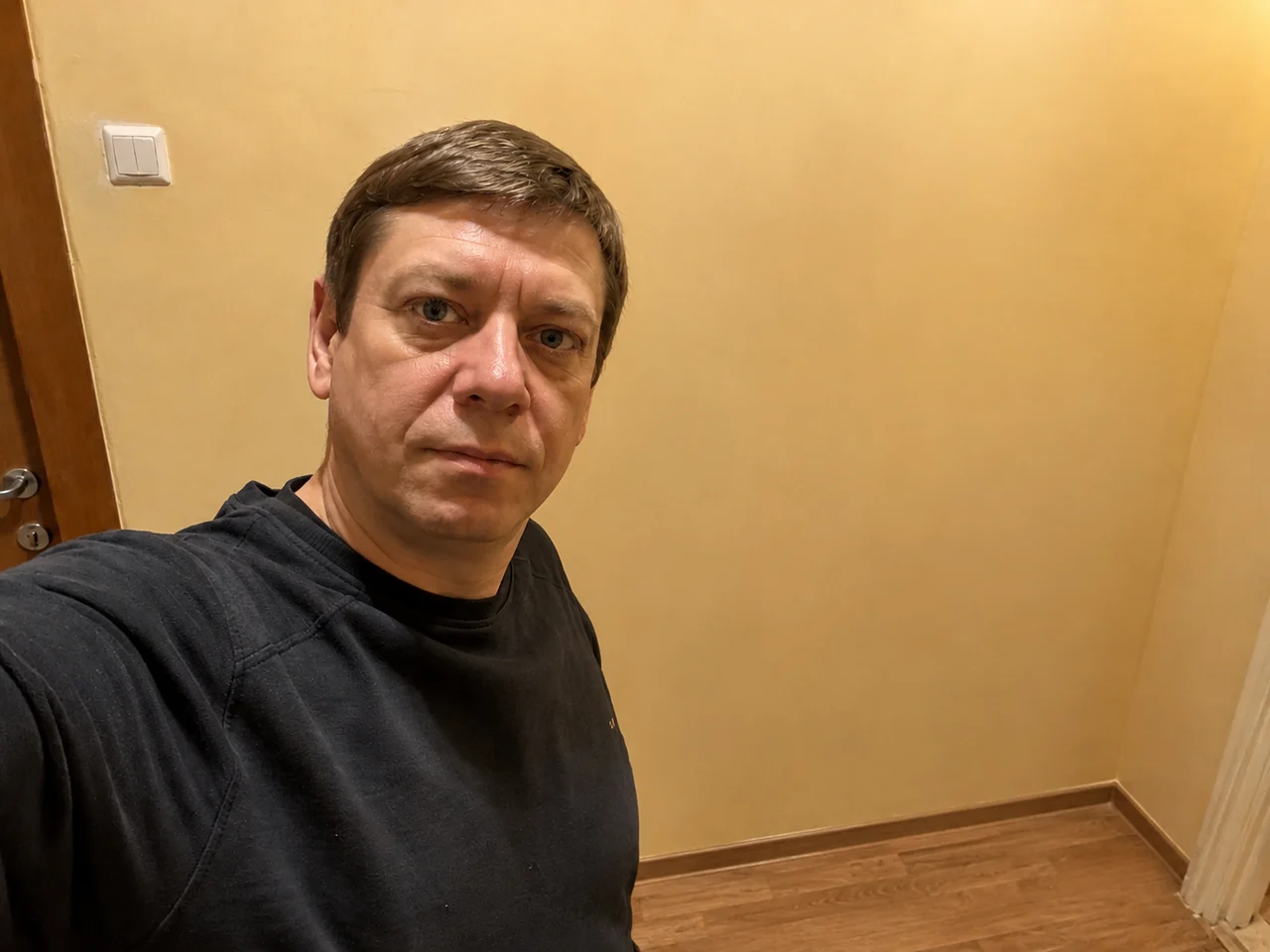 Мужчина в тёмном свитере в прихожей: вешалка с куртками, обувь и коробки убраны, за спиной осталась ровная стена